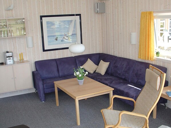 Una habitación con un sofá morado, una mesa de madera con un jarrón de flores, una silla, un cuadro en la pared y una ventana con cortinas amarillas.