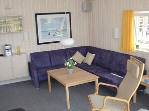 A room with a purple sofa, a wooden table with a vase of flowers, a chair, a framed picture on the wall, and a window with yellow curtains.