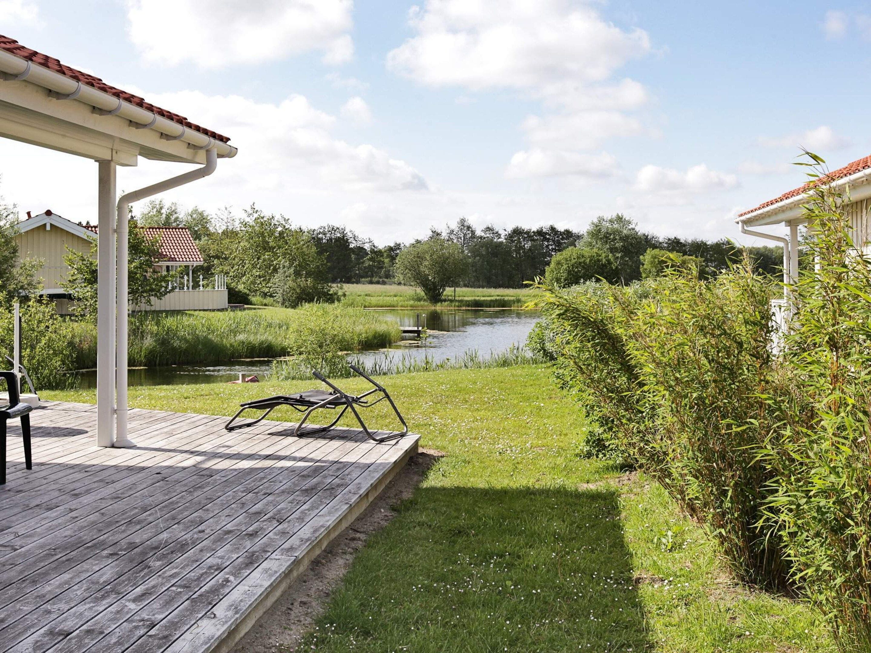 Una terraza de madera con una pérgola blanca, un estanque y follaje circundante.