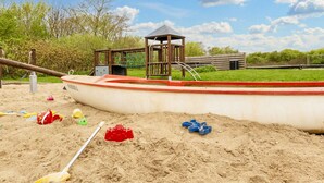 Un área de juegos con arena que presenta un castillo de arena con forma de barco, una estructura de madera y un pequeño refugio.