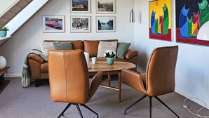 Uma sala de estar moderna com um sofá de couro marrom, uma mesa de centro de madeira e duas poltronas de couro. Há seis quadros na parede acima do sofá.