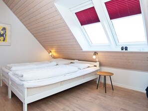 Una cama individual con cabecera de madera, una mesita redonda y un tragaluz con persianas rojas.