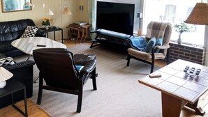 Una sala de estar con un sofá de cuero negro, una mesa de centro blanca, un sillón marrón y una mesa de madera con tazas. Un televisor está montado en la pared y hay una ventana con persianas.