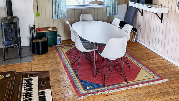 Una habitación acogedora con una mesa redonda, sillas blancas, un tapete con estampado rojo, un hogar de leña y un piano.