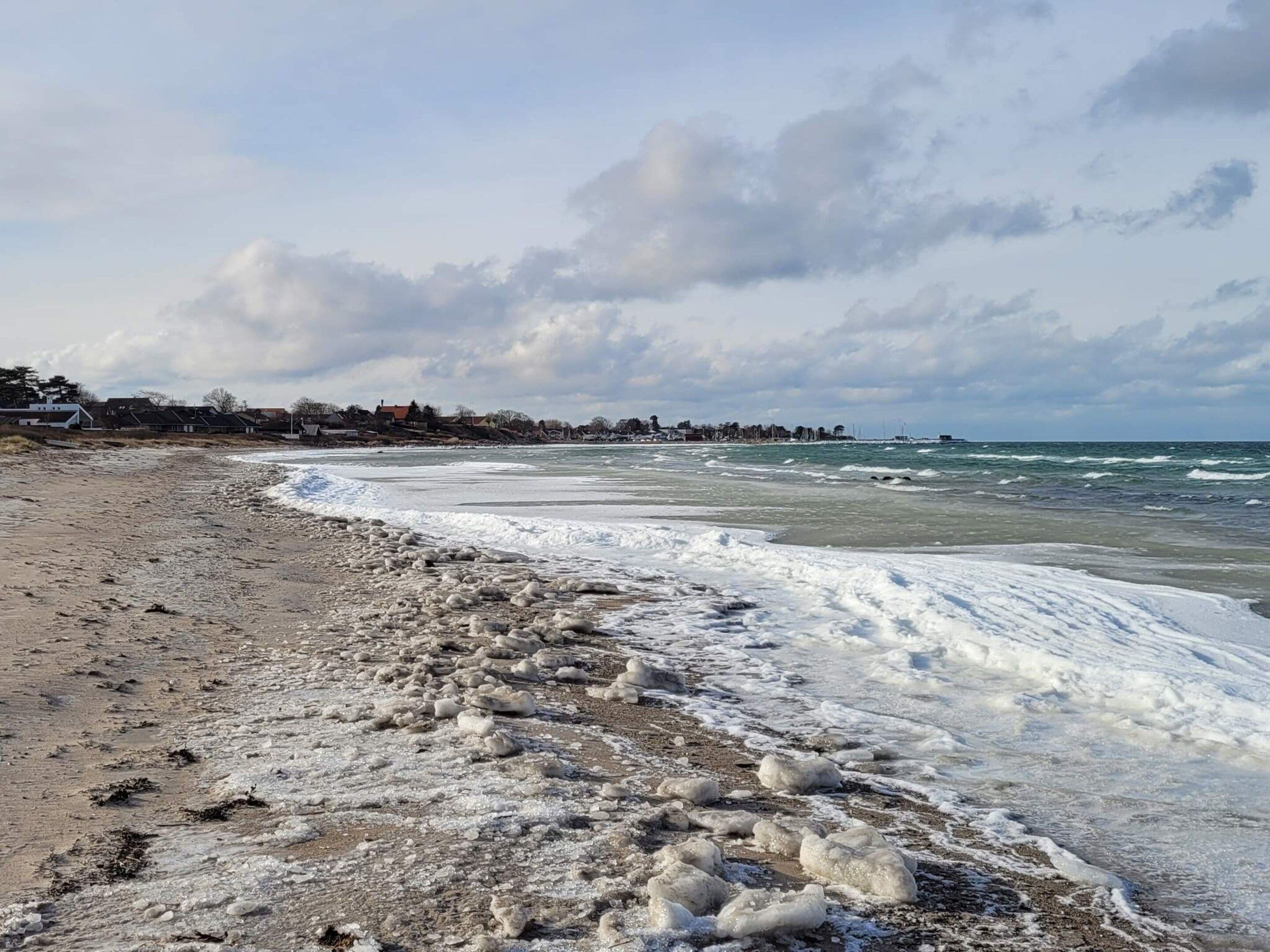 Une plage parsemée de formations de glace, avec des vagues et, au loin, une côte bordée de bâtiments.