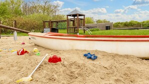 A sandy play area with a boat-shaped sandbox, a wooden play structure, and a small shelter. - 6 Person Holiday Home in Vestervig (Vestervig)