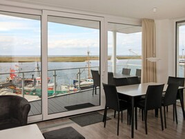 Una habitación moderna con un amplio ventanal corredera que da a un balcón con vistas al agua. En el interior, hay una mesa de comedor con sillas y un sofá.