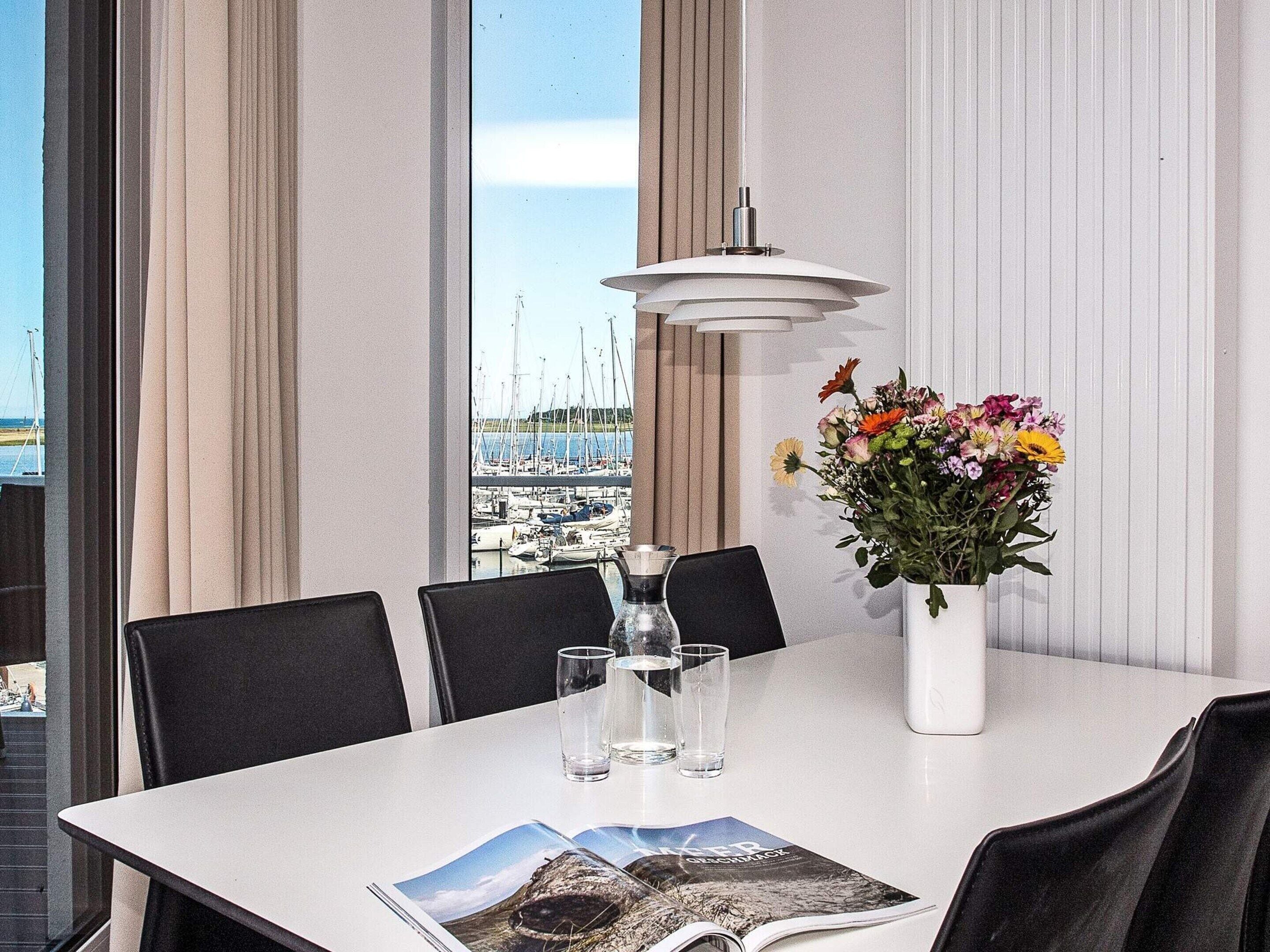 Un coin repas avec une table dressée pour deux personnes, un vase de fleurs et une vue sur une marina, visible à travers de grandes fenêtres.