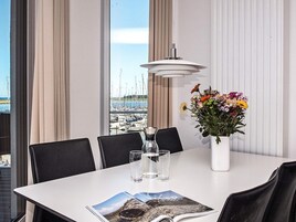 Un comedor con una mesa dispuesta para dos, un jarrón de flores y vistas a un puerto deportivo a través de amplias ventanas.
