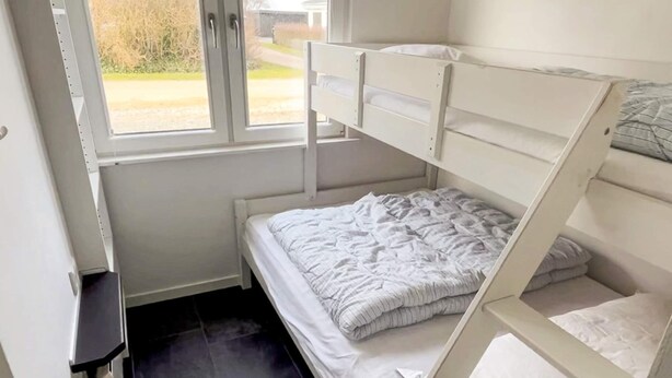 A bunk bed with a white headboard and footboard, a small bedside table, and a window with a view of a garden. - 4 Star Holiday Home in Vinderup (Vinderup)