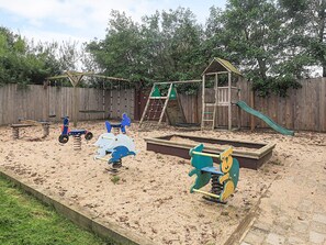 A sandy play area with various play structures, including a swing set, a climbing ladder, and a slide.