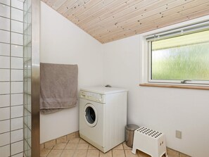 A compact laundry room with a washing machine, a drying rack, a trash bin, and a window with a view of greenery.