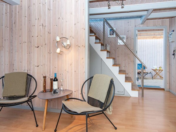 Un intérieur moderne avec un mur en bois, un escalier, deux fauteuils en osier, une petite table avec une bouteille et un verre, et une vue sur un balcon.