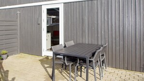 Un patio con una mesa y sillas, una puerta que da a una habitación con estantes, y una planta pequeña en maceta.