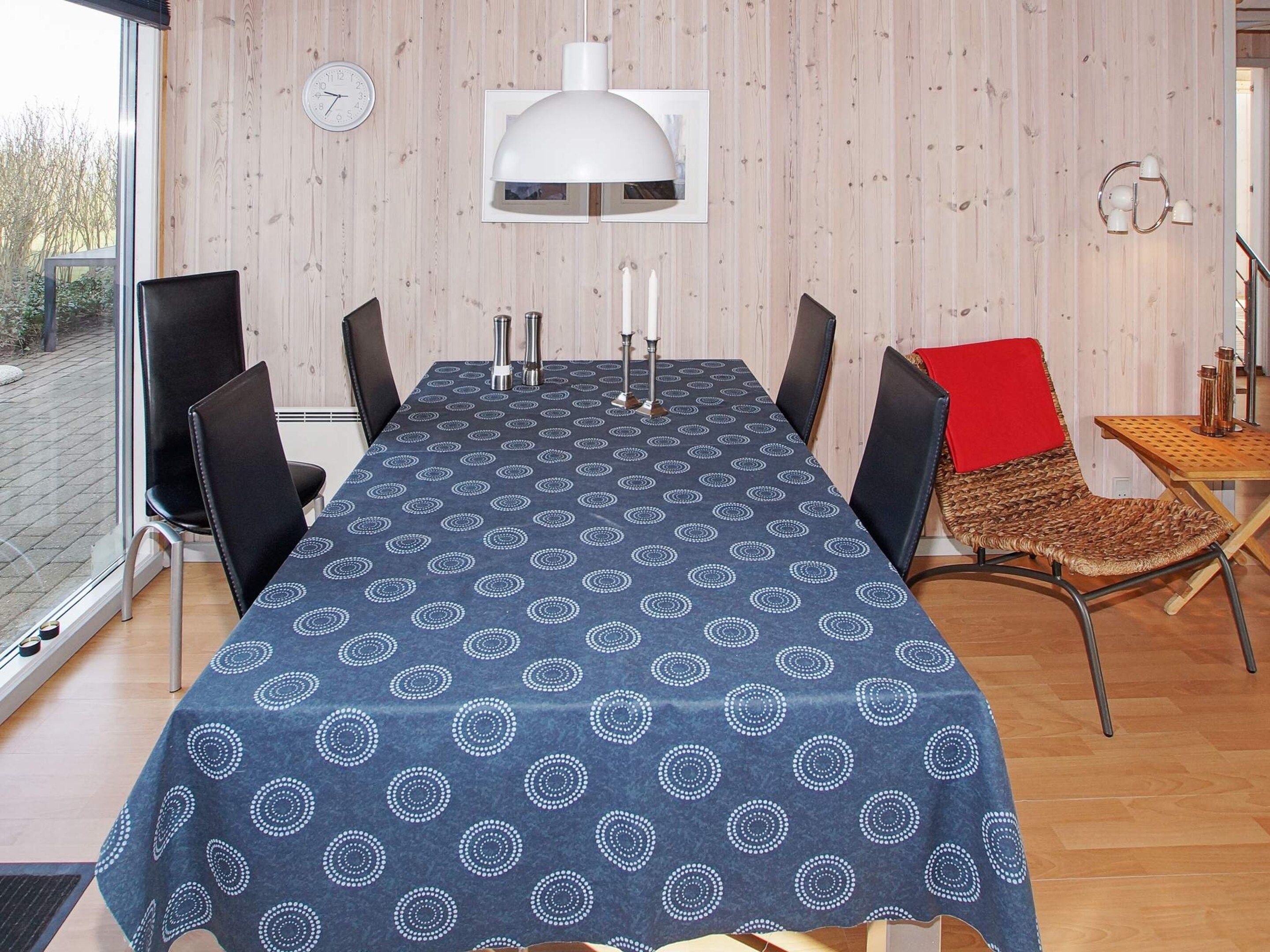 Un coin repas avec une table dressée pour quatre personnes, entourée de chaises, une horloge au mur et une fenêtre donnant sur l’extérieur.