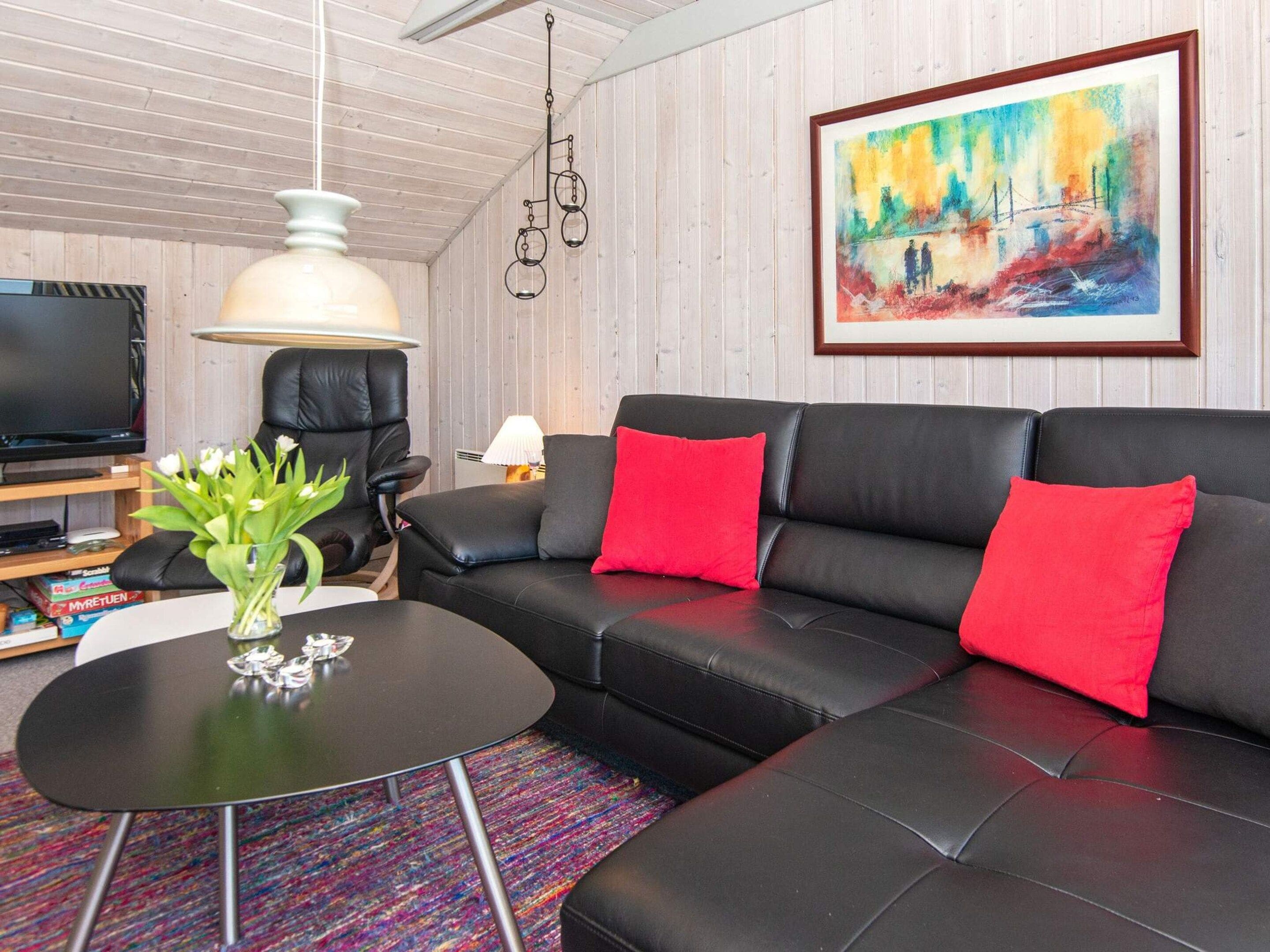 En moderne stue med en sort lædersofa, røde puder, et rundt sofabord, en hvid vase med blomster, et stort maleri på væggen og et fjernsyn på et træmøbel.