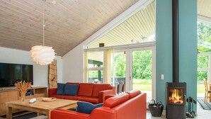 A spacious living room with a red sofa, wooden coffee table, and a TV. There's a fireplace and a large window overlooking greenery. - 8 Person Holiday Home in Brovst (Brovst)