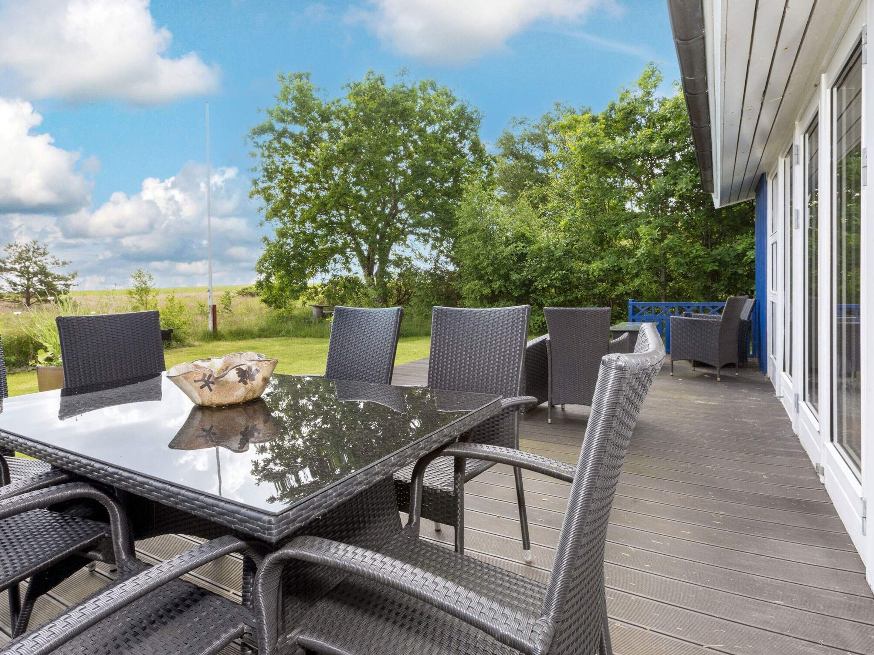 Une terrasse extérieure avec une table en verre et des chaises en osier, entourée d’une plateforme en bois et offrant une vue sur la verdure.
