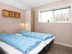 A room with two beds, a window with a view of trees, a shelf with a jar, and a wall-mounted light.