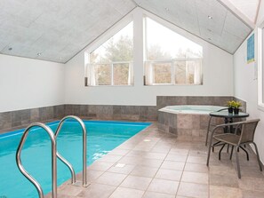 An indoor swimming pool with a hot tub, a table with chairs, and large windows.
