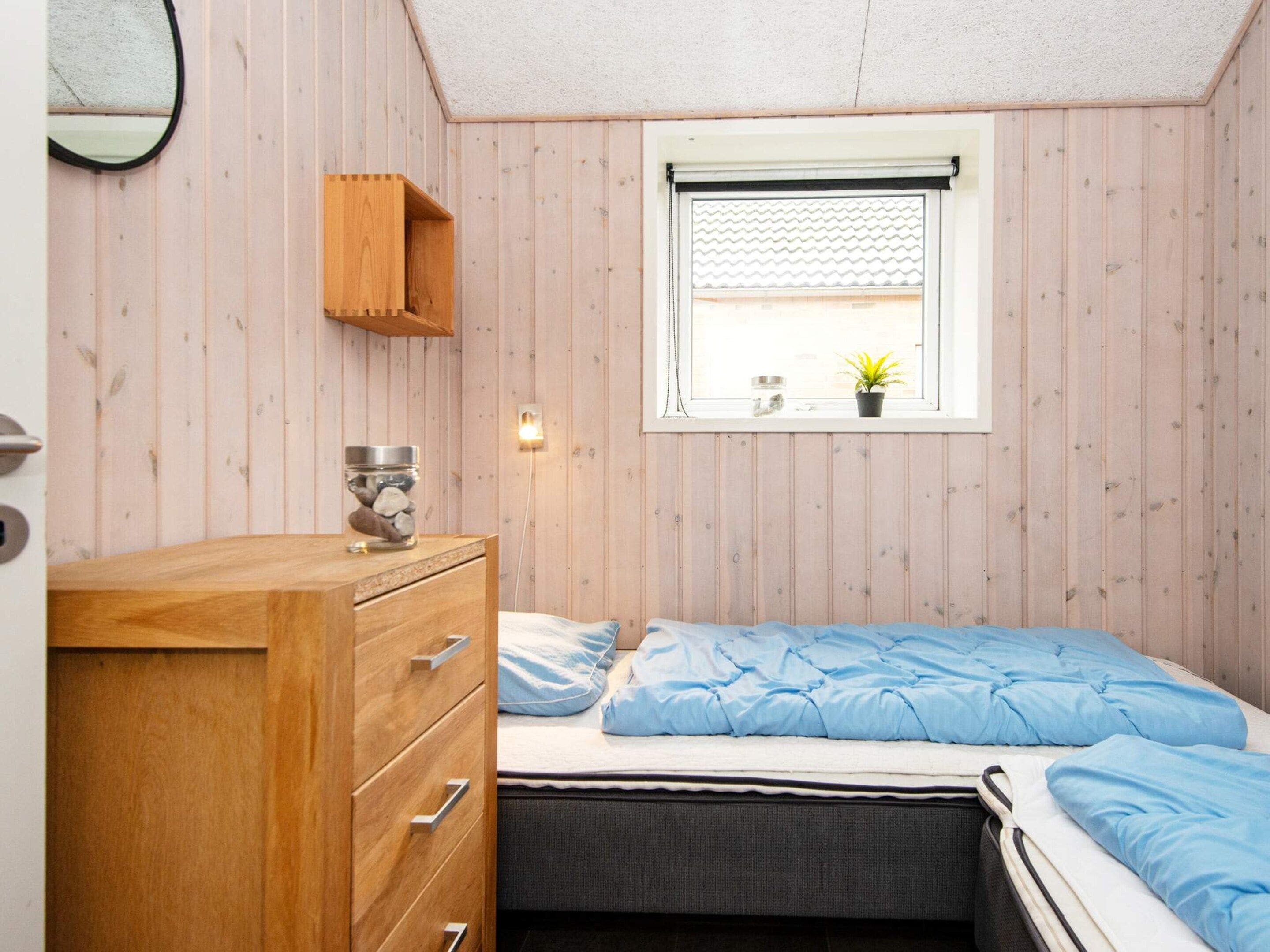 A compact bedroom with a single bed, a wooden dresser, a small window with a plant, and a wall-mounted shelf.
