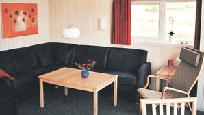 Una habitación con un sofá de color oscuro, una mesa de centro de madera con un jarrón, una silla, una mesita con una vela y una ventana con cortinas.