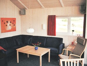 Une pièce avec un canapé de couleur sombre, une table basse en bois sur laquelle se trouve un vase, un fauteuil, une petite table avec une bougie et une fenêtre avec des rideaux.