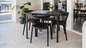 An outdoor dining area with a round table and black chairs, surrounded by potted plants and a tiled floor.