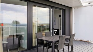 A modern outdoor dining area with a table and chairs, a glass door leading to a balcony, and a view of the sky and distant landscape.