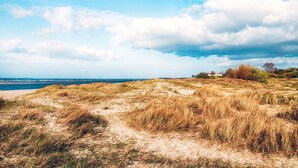 A coastal landscape with a sandy path leading to the sea, grassy dunes, and a distant building. - Holiday Home in Wendtorf (Schönberg)