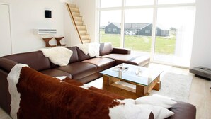 A modern living room with a brown leather sofa, a glass coffee table, and a large window offering a view of a house and greenery. - 5 Star Holiday Home in Lokken (Lokken)