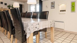 A dining area with a long table, black chairs, and a white tablecloth. There are two pendant lights, an air conditioning unit, and a window with blinds. - 9 Person Holiday Home in Blavand (Blavand)