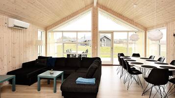 A spacious room with a large wooden ceiling, a sectional sofa, a dining table with chairs, and large windows offering a view of the outdoors.