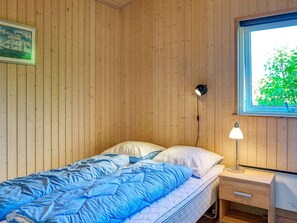 A single bed with a blue comforter and white pillows, a bedside table with a lamp, and a window with a view of greenery. - 4 Star Holiday Home in Gilleleje (Gilleleje)