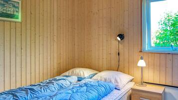 En enkeltseng med et blåt dynebetræk og hvide puder, et sengebord med en lampe, et billede i ramme på væggen og et vindue med udsigt til grønne omgivelser.