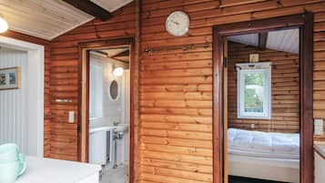 Interior de una cabaña de madera con baño a la vista, una cama y un reloj en la pared.