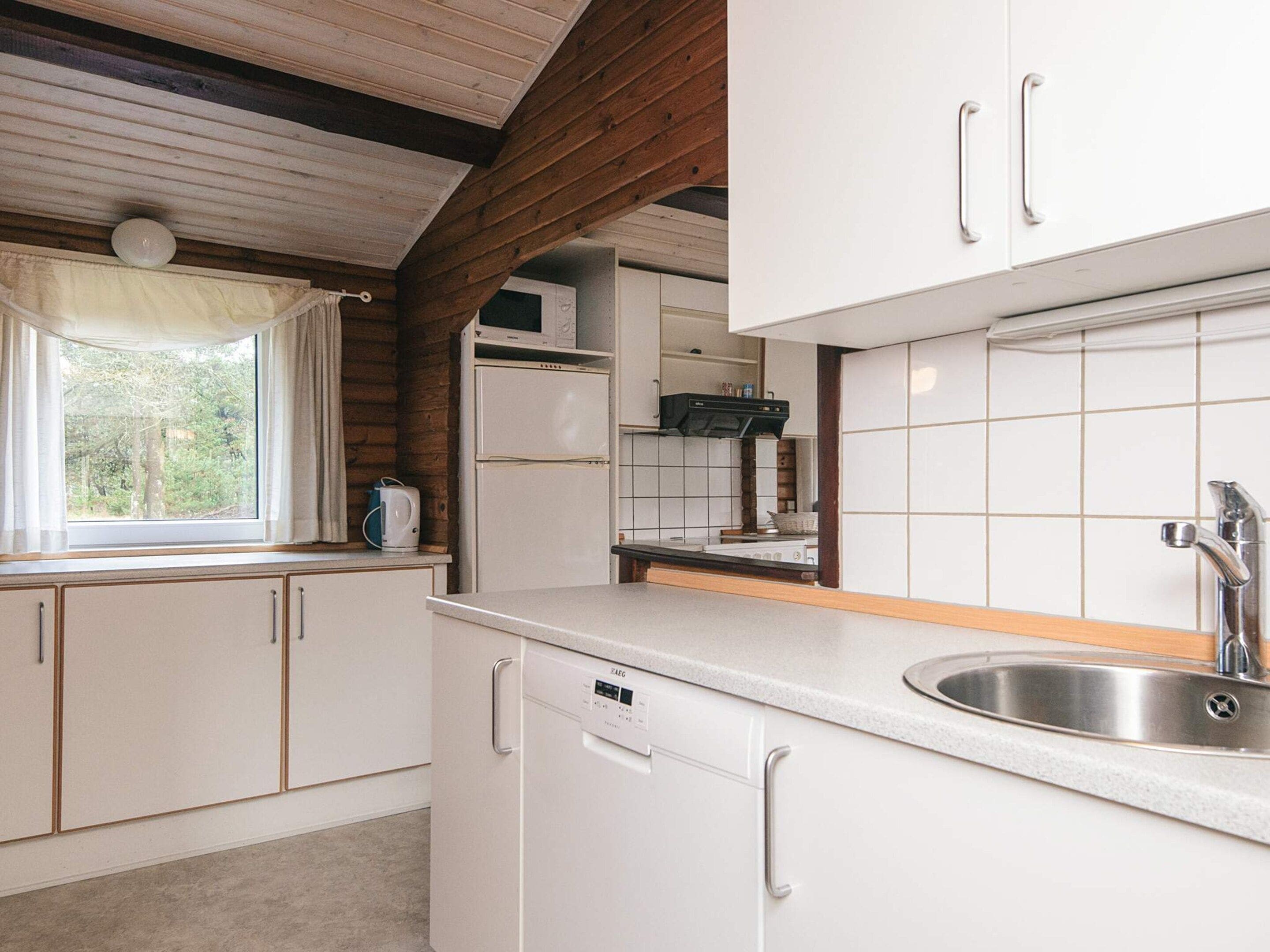 Una cocina pequeña con gabinetes blancos, un lavamanos de acero inoxidable y un ventanal con cortinas.