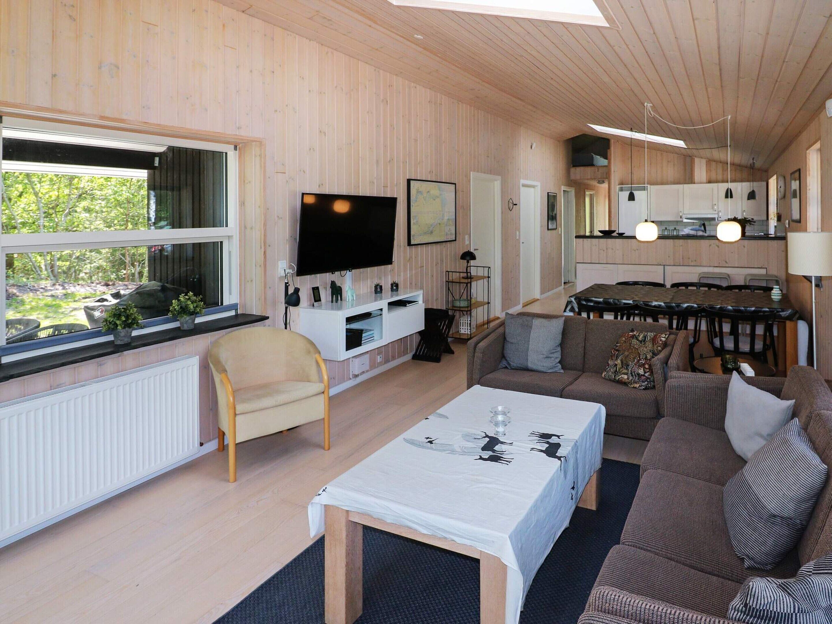 Een moderne woonkamer met een bank, een salontafel en een televisie. Er is een eethoek met een tafel en stoelen. De kamer heeft houten wanden en een groot raam.