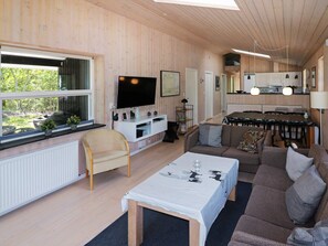 A modern living room with a sofa, a coffee table, and a TV. There is a dining area with a table and chairs. The room has wooden walls and a large window.