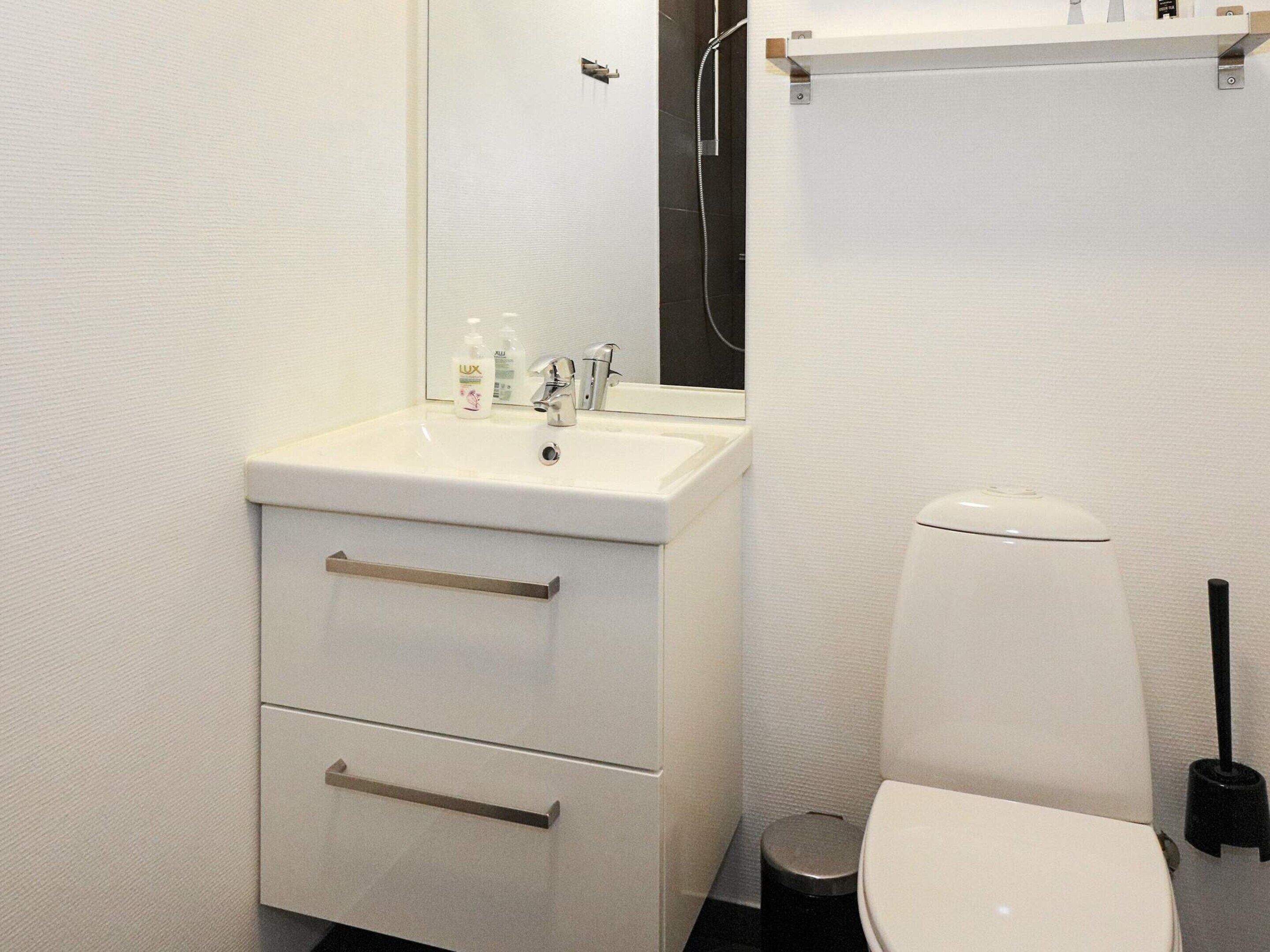 Un baño con lavamanos blanco, espejo e inodoro. Hay dos cajones debajo del lavamanos y un pequeño cubo de basura junto al inodoro.