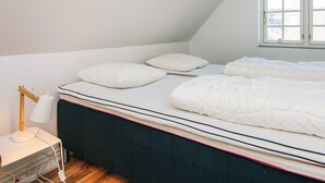 Un dormitorio con una cama individual, ropa de cama blanca, una mesita de noche con lámpara y una ventana con persianas.