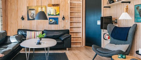 A modern living room with a black leather sofa, a round coffee table, and a grey armchair. There are wooden walls and a ceiling with exposed beams.