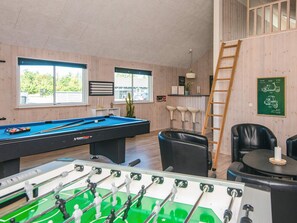 A pool table, foosball table, and bar area in a room with wooden walls and windows.
