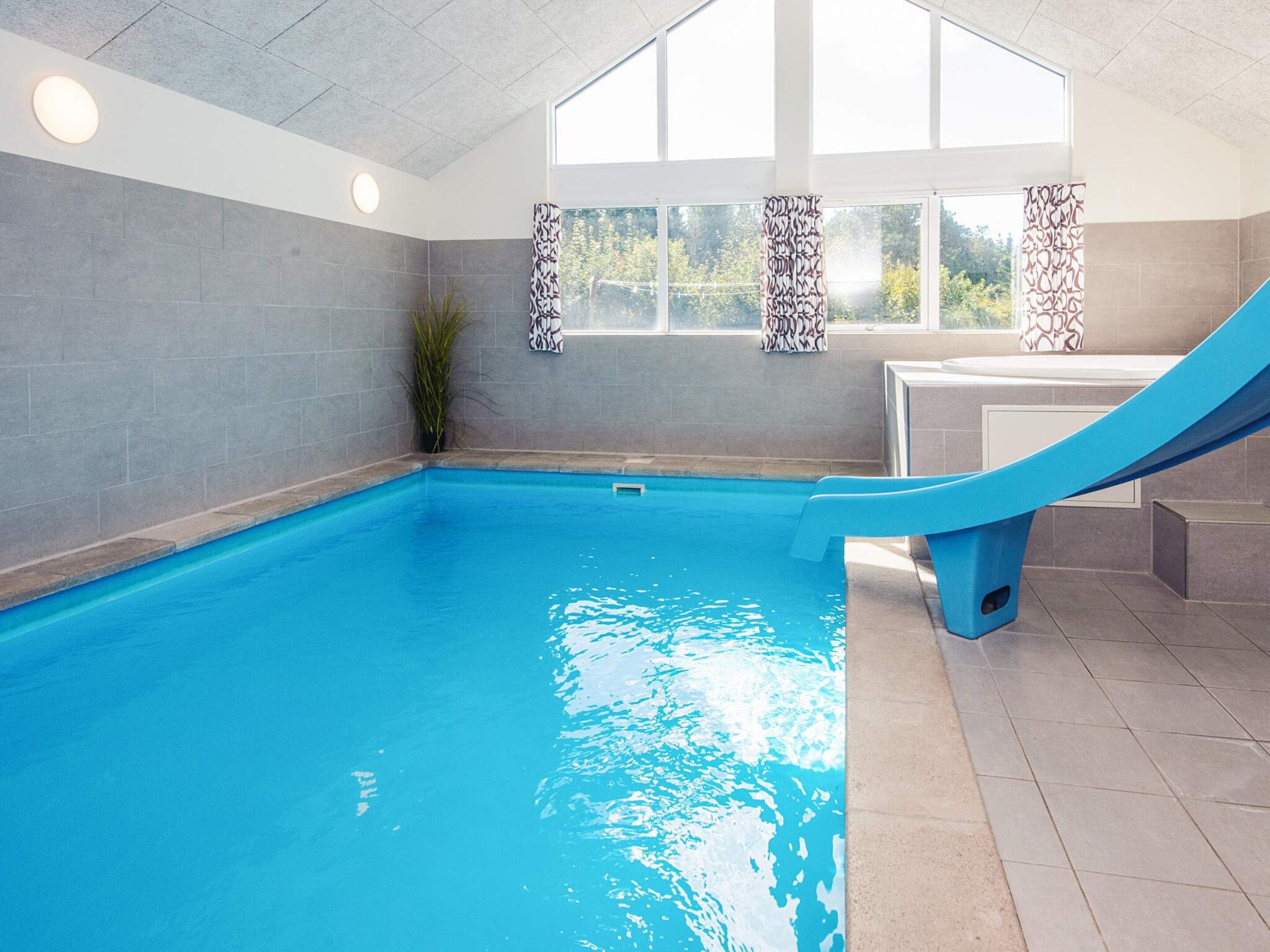 Ein Hallenbad mit blauer Rutsche, grauen Fliesenwänden und einem Fenster mit gemusterten Vorhängen.