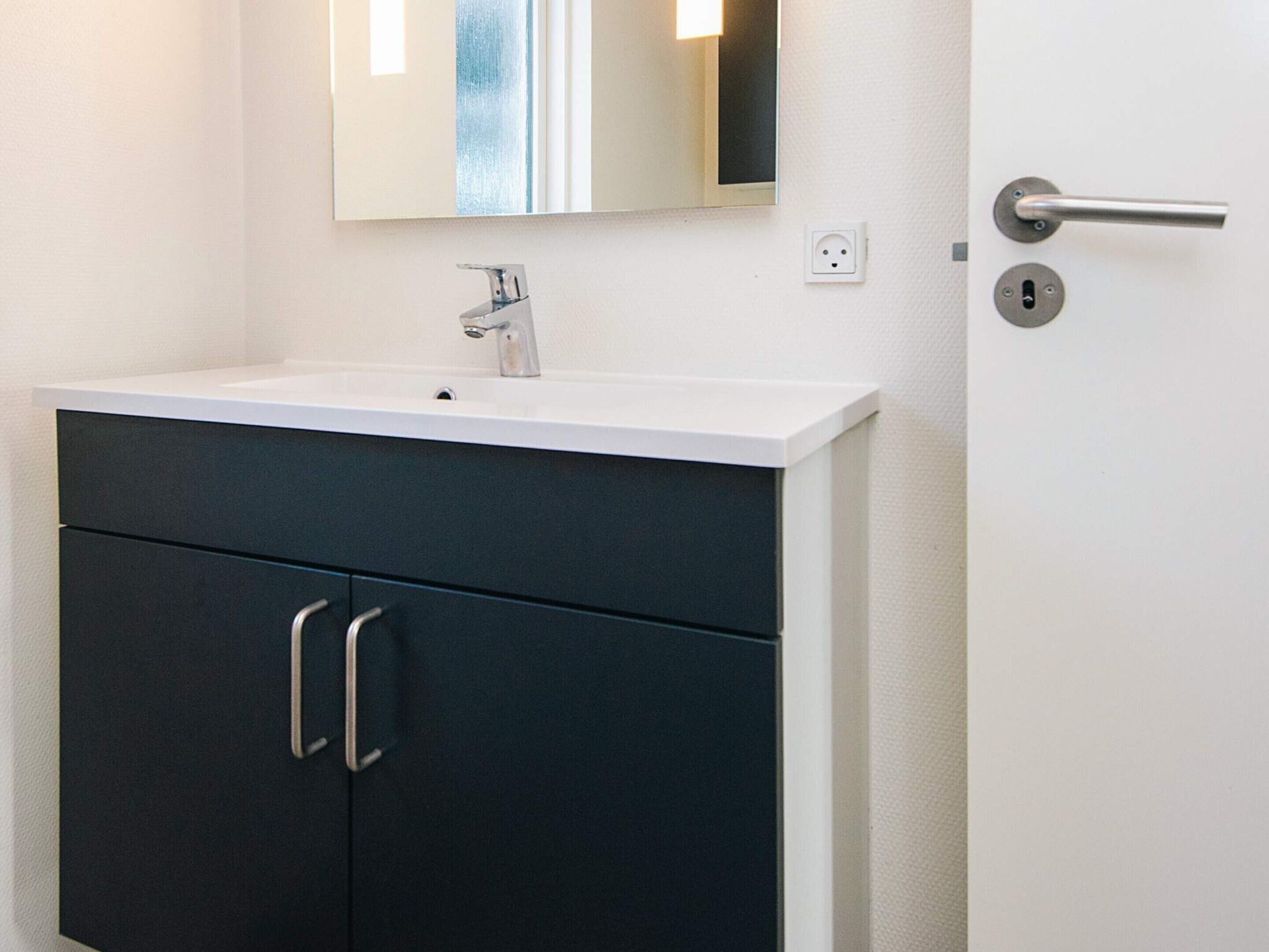Une salle de bain avec un lavabo blanc, des meubles sombres, un miroir et une porte dotée d’une poignée.
