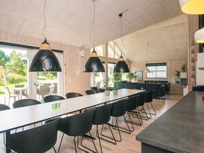 A modern dining area with a long table, black chairs, pendant lighting, and a view of the outdoors.