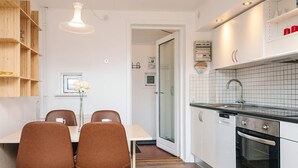 A modern kitchen with white cabinets, a black countertop, and stainless steel appliances. There is a dining area with a table and chairs, and a wooden shelf unit. - 6 Person Holiday Home in Romo (Rømø)