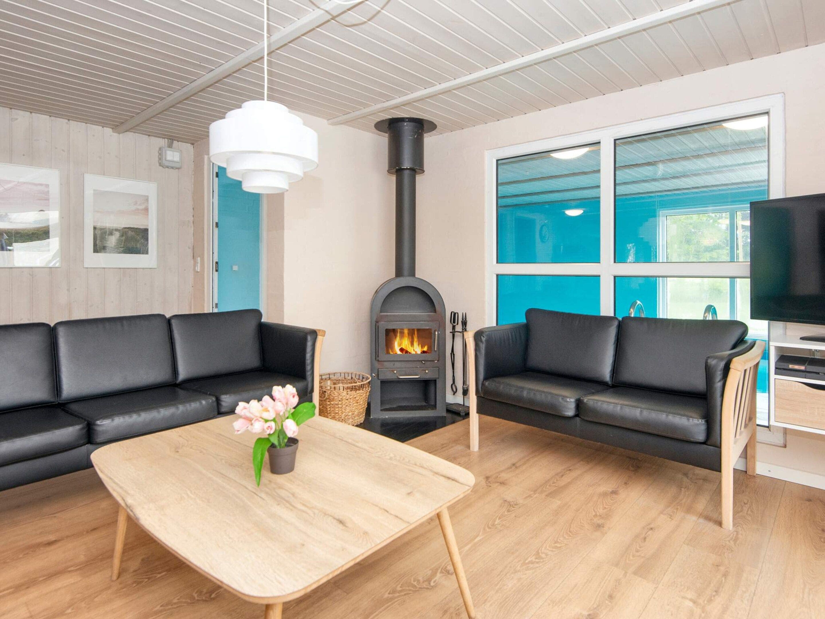 A modern living room with two black leather sofas, a wooden coffee table with a vase of flowers, a fireplace, and a large window with blue-tinted glass.