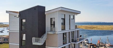 Un edificio moderno de varios pisos con balcón, ventanas amplias y vista a un puerto deportivo con barcos.