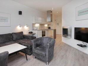 A modern living room with a grey sofa, a small armchair, a TV, and a kitchen area with cabinets and a microwave.